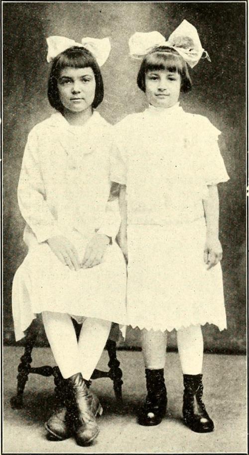 Two young girls in white dresses and large bows, seated side by side, with vintage-style boots and a neutral background.