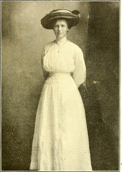 A woman in a white dress and large hat stands confidently, posing for a vintage photograph.