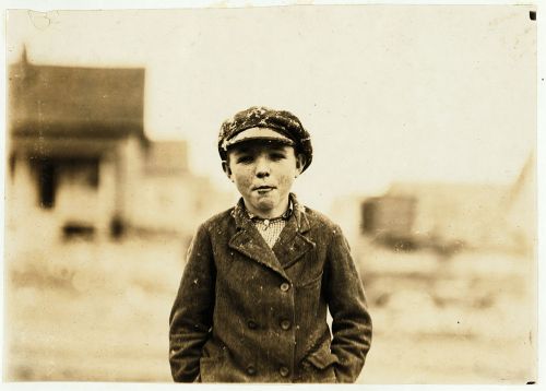 A young boy in a cap and coat stands outdoors, with a blurred background of buildings and a dusty landscape.