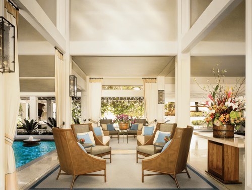 A stylish lounge area with wooden chairs, colorful cushions, and a view of a pool and tropical plants.