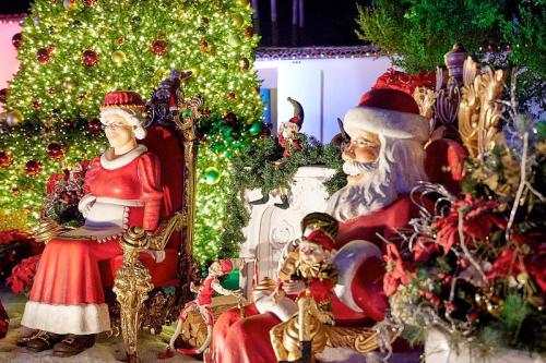 A festive scene featuring Santa and Mrs. Claus sitting on ornate chairs, surrounded by Christmas decorations and a tree.