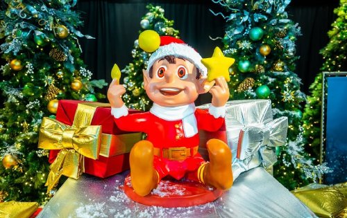 A cheerful elf in a Santa hat sits among Christmas trees and presents, holding a star and a candle.