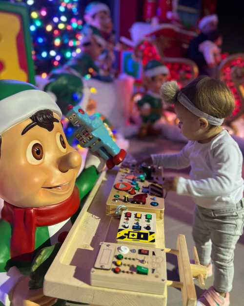 A child interacts with a colorful toy workstation beside a large, cheerful elf figure in a festive setting.