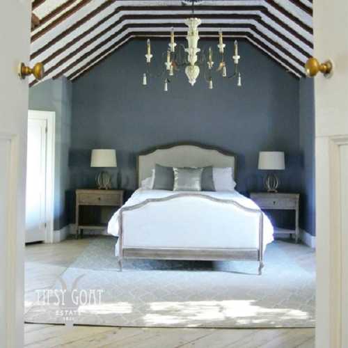 A stylish bedroom with a gray accent wall, chandelier, and elegant furnishings, featuring a cozy bed and lamps.