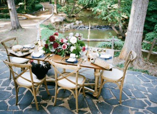 A beautifully set outdoor dining table with flowers, plates, and a rustic backdrop of trees and a stream.