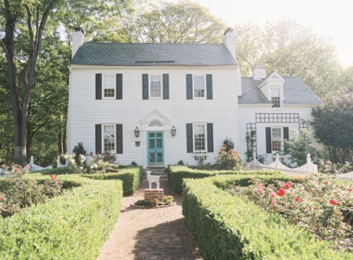 A charming white house with black shutters, surrounded by manicured hedges and blooming rose bushes in a sunny garden.