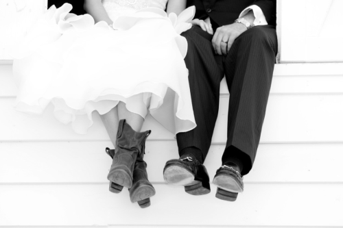 A close-up of a bride in a white dress and a groom in a suit, sitting together with their feet hanging off a ledge.