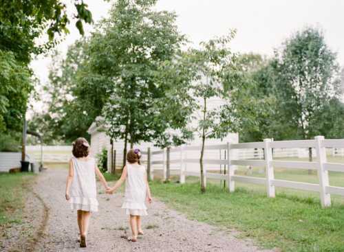 Two girls in white dresses walk hand in hand down a gravel path, surrounded by greenery and a white fence.