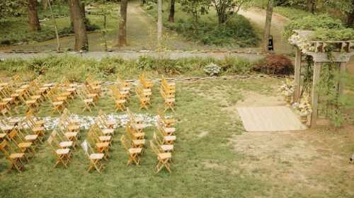 An outdoor wedding setup with wooden chairs arranged in rows and a floral-decorated archway in a lush garden.