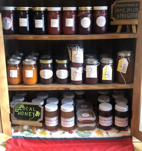 A wooden shelf displaying various jars of homemade jams, jellies, preserves, and local honey.