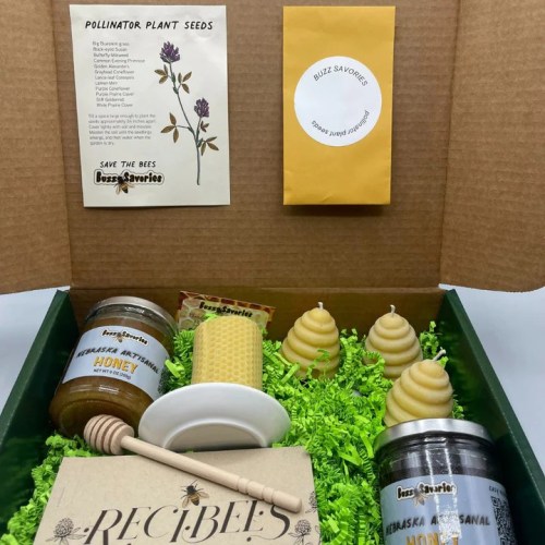 A gift box containing pollinator plant seeds, honey jars, beeswax candles, and a honey dipper on green filler.