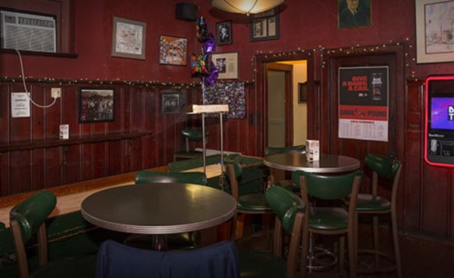 Cozy bar interior with green chairs, wooden walls, and framed artwork, featuring a small table and dim lighting.