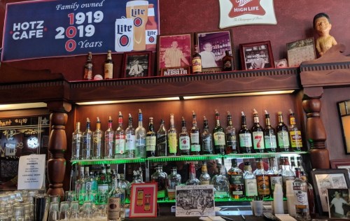 A bar shelf displaying various liquor bottles, framed photos, and a sign celebrating 100 years of family ownership.