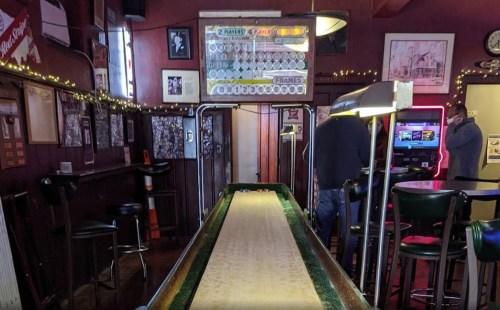 A shuffleboard table in a dimly lit bar, with a scoreboard and patrons in the background.