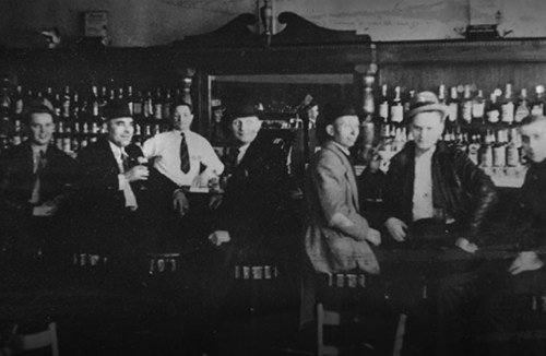 A black-and-white photo of seven men sitting at a bar, surrounded by bottles on shelves in a vintage setting.