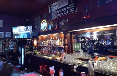 A cozy bar interior with a well-stocked shelf of liquor, a TV, and festive decorations. A bartender is visible.