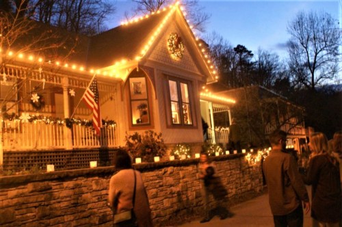A cozy house decorated with holiday lights, surrounded by people walking along a path at dusk.
