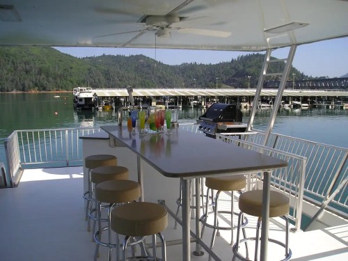 A spacious boat deck with a bar, colorful drinks, and a view of a marina and green hills in the background.