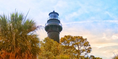 A tall lighthouse surrounded by palm trees and greenery under a blue sky with wispy clouds.