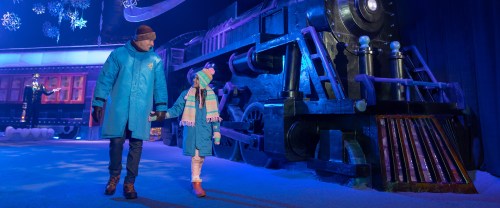 A man and a child in winter coats walk hand in hand near a large, colorful train set in a snowy scene.