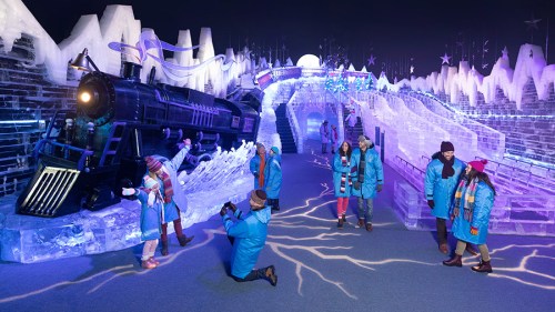 Visitors in blue jackets explore an ice-themed attraction featuring an ice train and colorful ice sculptures.