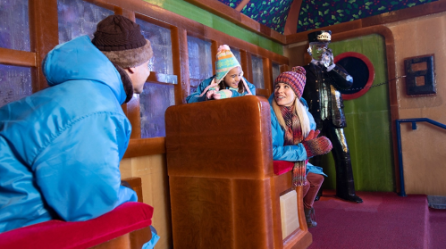 Two people in winter coats chat happily while a third person sits nearby, all in a colorful, cozy indoor setting.