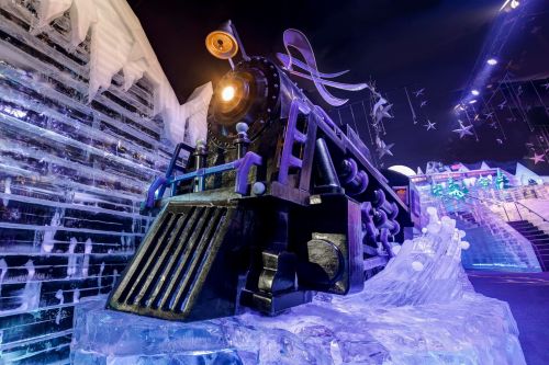A large ice sculpture of a steam locomotive, illuminated with colorful lights, surrounded by ice formations.