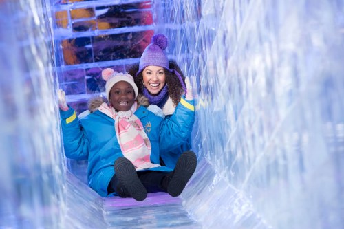Two women, one adult and one child, smile while sitting on a colorful ice slide surrounded by ice sculptures.