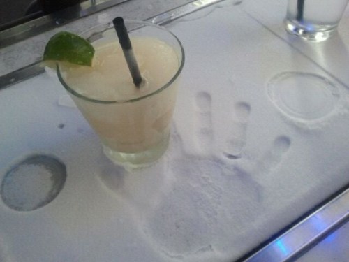 A cocktail with a lime wedge sits on a snowy surface, next to a handprint and circular impressions.
