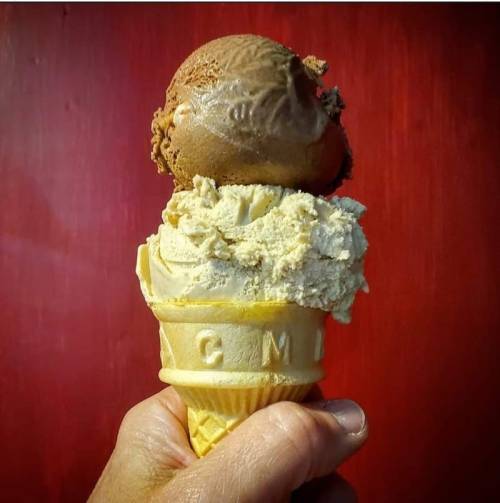 A hand holding a double scoop ice cream cone with chocolate and a light-colored flavor against a red background.