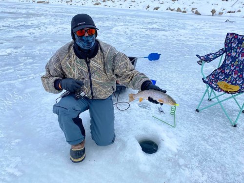 A person in winter gear kneels on ice, holding a fish near an ice fishing hole, with a chair in the background.