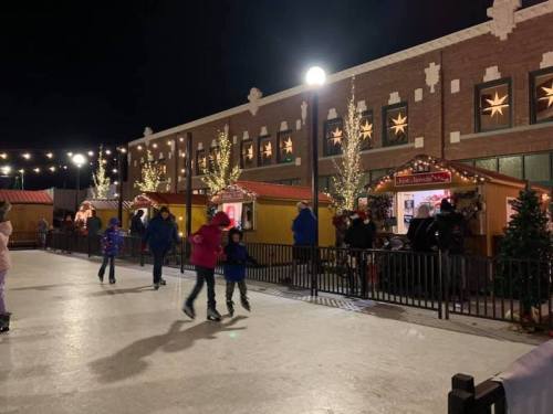 A nighttime ice skating rink with people skating and festive decorations on nearby buildings.