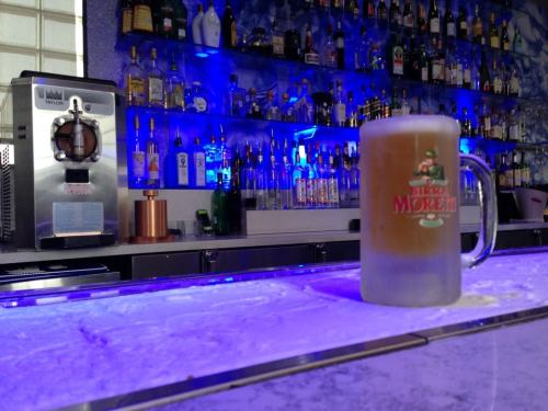 A frosty mug of beer sits on a bar counter, with a colorful backlit shelf of liquor bottles in the background.