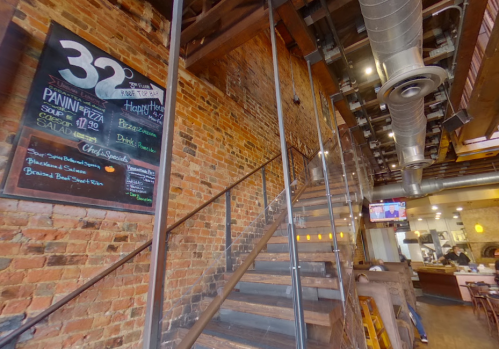 Interior of a restaurant featuring a brick wall, a chalkboard menu, and a staircase leading to an upper level.