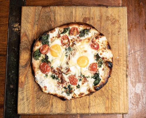 A freshly baked pizza topped with spinach, cherry tomatoes, eggs, and cheese on a wooden cutting board.
