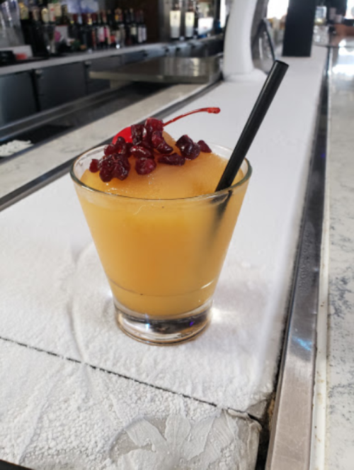 A frozen cocktail in a glass, topped with cherries, sits on a bar counter covered in ice.
