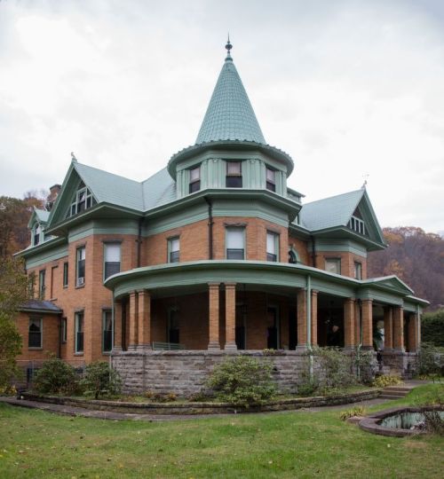 A large, historic brick house with a green roof and turret, surrounded by gardens and trees under a cloudy sky.