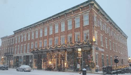 Historic brick building adorned with holiday decorations, surrounded by falling snow and a wintery street scene.