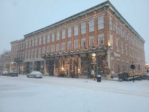 A historic brick building decorated for the holidays, surrounded by falling snow and a snowy street.