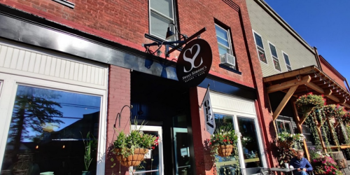 Exterior of a café with a sign reading "S2," featuring large windows and hanging flower baskets. Bright blue sky above.