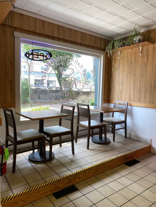A cozy cafe interior with empty tables and chairs, featuring a large window and wooden paneling. A "Open" sign is visible.