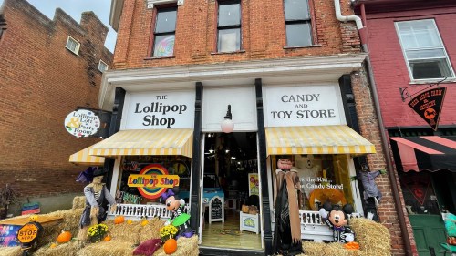A colorful storefront of "The Lollipop Shop," featuring candy, toys, and autumn decorations with hay bales outside.