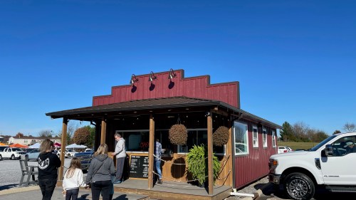 A small red building with a porch, people approaching it under a clear blue sky, and a white truck parked nearby.