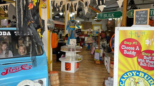 A colorful store interior filled with decorations, candy displays, and a vintage blue soda truck. Kids are visible inside the truck.