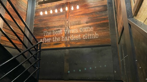 A staircase with a wooden wall featuring the quote, "The best views come after the hardest climb," illuminated by hanging lights.