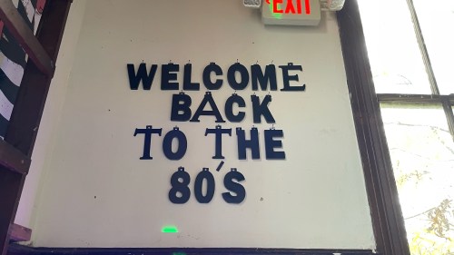 Sign on a wall reads "WELCOME BACK TO THE 80'S" in bold letters, with an exit sign above.