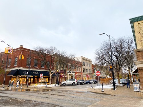 A street view of a small town with shops, trees, and a cloudy sky, showing pedestrians and parked cars.