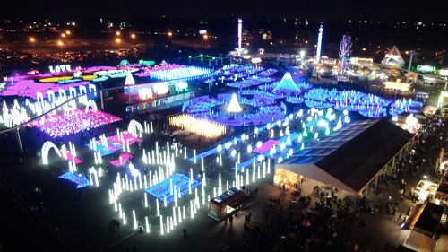 Aerial view of a vibrant night festival with colorful lights, attractions, and a large crowd enjoying the festivities.