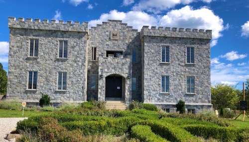 A stone castle with a symmetrical facade, featuring turrets and a landscaped garden in front under a blue sky.