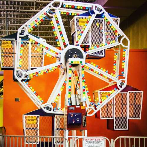 A colorful indoor Ferris wheel with bright lights and enclosed cabins against an orange wall.
